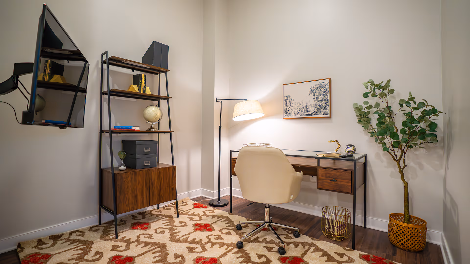A cozy home office setup featuring a wooden desk with drawers, a beige swivel chair, a floor lamp, a framed black and white landscape artwork on the wall, a tall shelving unit with books and decorative items, a potted plant in a woven basket, and a patterned area rug on a wooden floor.