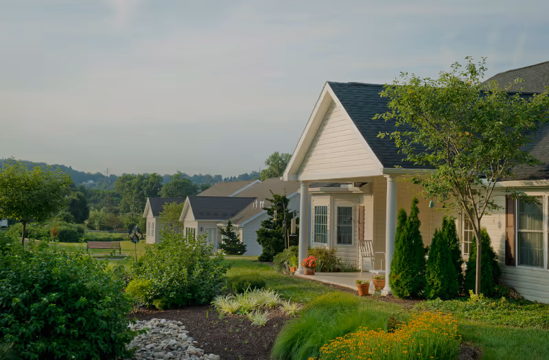 View of a senior living community with several single-story houses surrounded by well-maintained gardens, green lawns, and trees under a clear sky.