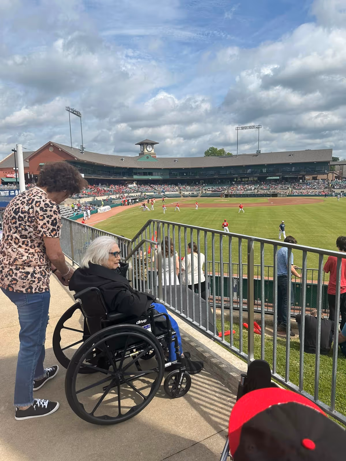 An elderly woman in a wheelchair, accompanied by another woman, is watching a baseball game at a stadium. The field and players are visible in the background under a partly cloudy sky.