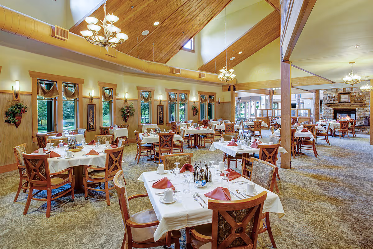 A spacious dining room with multiple tables set with white tablecloths, red napkins, cups, and silverware. The room features large windows with wooden frames, decorative wall hangings, and plants. There are chandeliers hanging from a high wooden ceiling, and a stone fireplace is visible in the background.