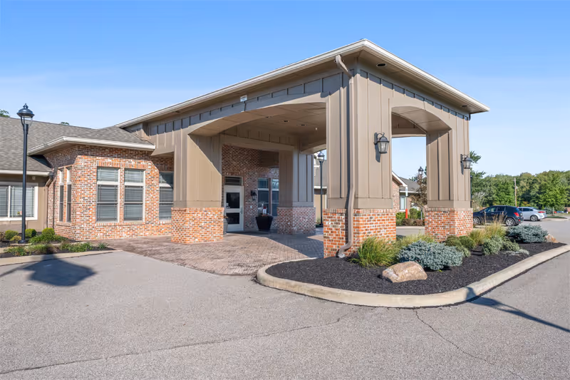Entrance of Cardinal Court Assisted Living & Memory Care facility showing a covered drop-off area with brick and beige panel exterior, surrounded by landscaped plants and a parking lot with cars in the background under a clear blue sky.