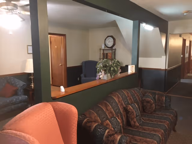 Interior view of a senior living facility lounge area with patterned sofas and armchairs, a wooden shelf with a clock and plants, and a hallway with doors leading to other rooms.