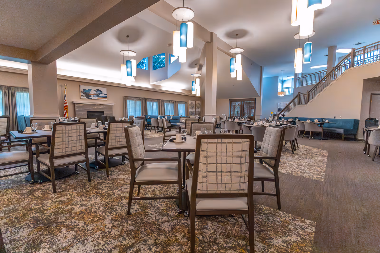 Spacious dining room in a senior living facility with multiple tables and chairs arranged neatly. The room features high ceilings with modern hanging light fixtures, large windows with curtains, a fireplace with an American flag beside it, and a staircase leading to an upper level. The carpet has a patterned design and the tables are set with cups and glasses.