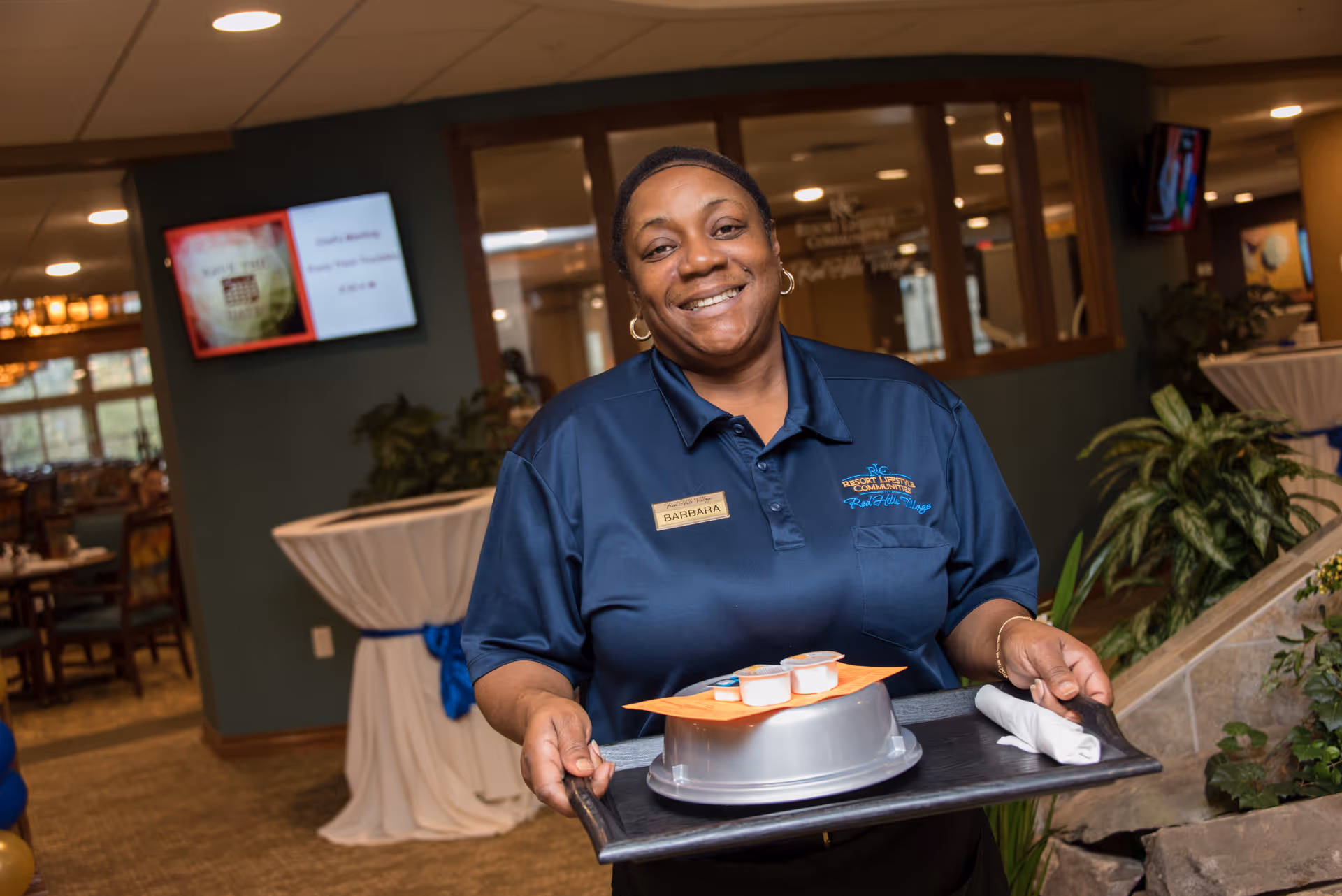 A smiling staff member in a navy polo carries a covered meal tray in a retirement facility dining area.