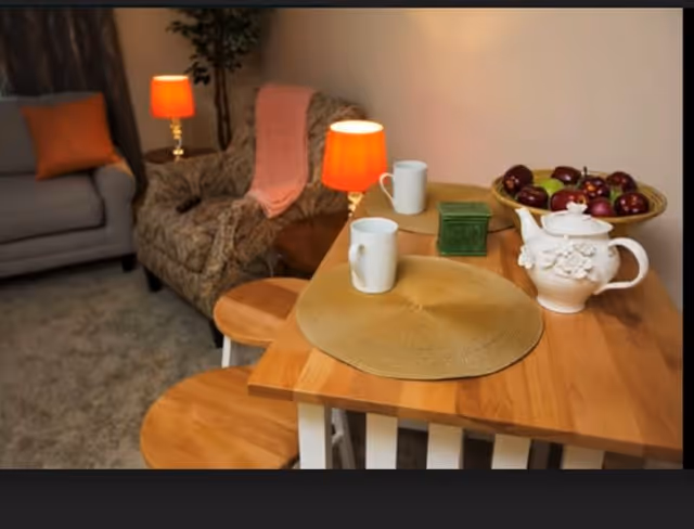 A cozy living area with a wooden table set with two placemats, two white mugs, a decorative white teapot, a green container, and a bowl of red apples. In the background, there is a patterned armchair with a pink throw blanket, a gray sofa with an orange pillow, two small table lamps with orange shades, and a potted plant.