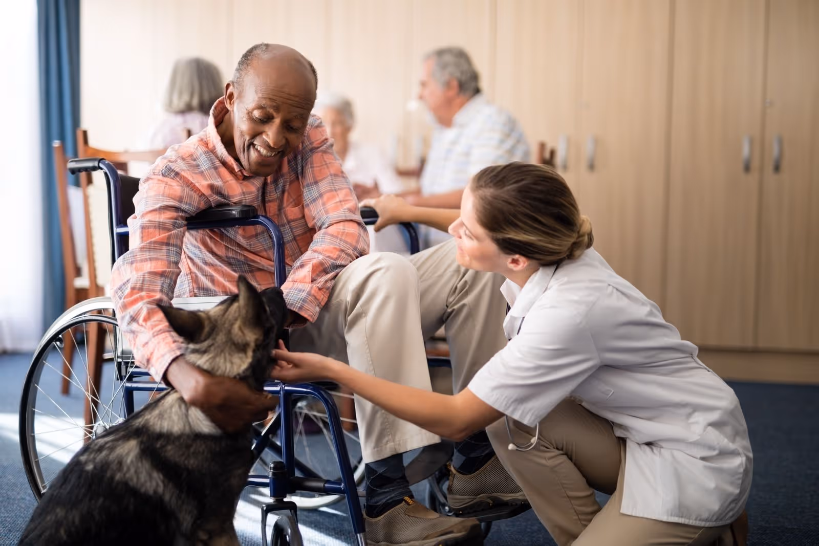 A smiling elderly man in a wheelchair petting a large dog while a female caregiver kneels beside him, also interacting with the dog. In the background, other elderly individuals are seated and engaged in conversation in a well-lit room.