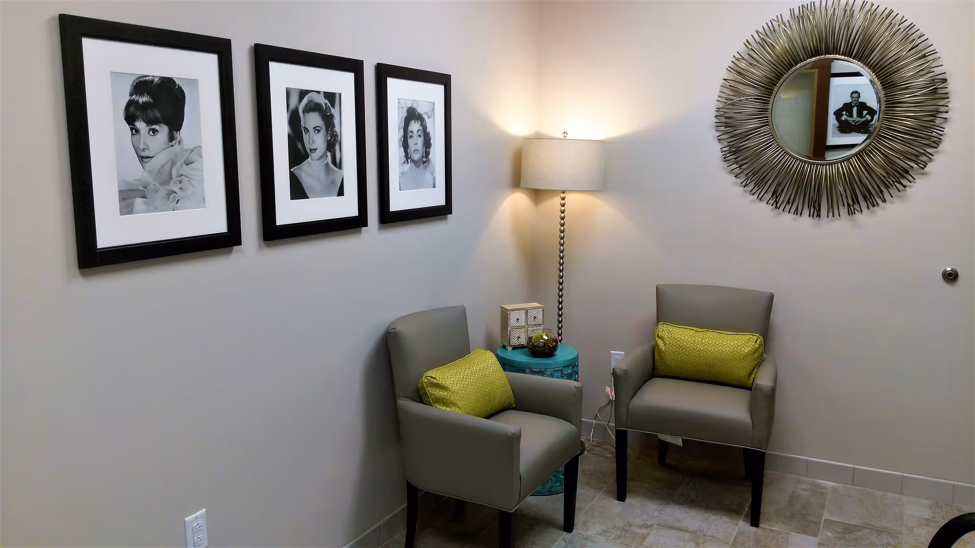 Small seating area with two gray armchairs with green pillows, a floor lamp, a round decorative mirror and three framed black-and-white portraits on a beige wall.
