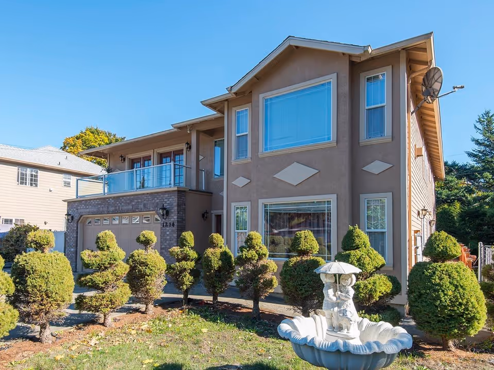Exterior view of a two-story residential building with beige walls and multiple windows. The front yard features neatly trimmed topiary bushes and a white decorative fountain with a statue of two children holding an umbrella. The sky is clear and blue.