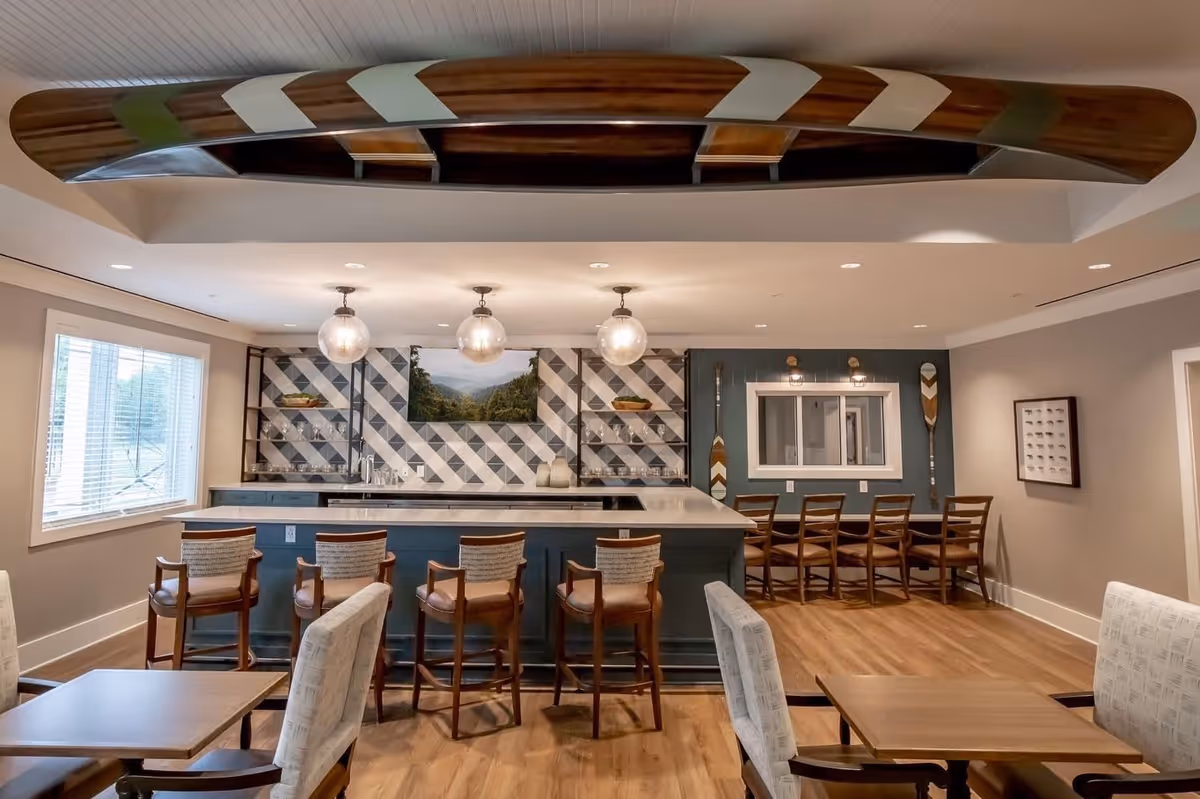 Interior view of a communal dining and bar area with wooden floors, a bar counter with six high chairs, and additional seating with tables and chairs. The back wall features a geometric tile pattern with shelves and a landscape picture. Three globe pendant lights hang above the bar, and decorative wooden paddles are mounted on the walls. A large wooden canoe is mounted on the ceiling.
