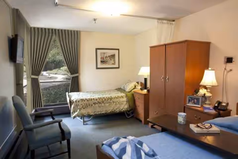 Bedroom in a senior living facility with bed, wardrobe, dresser, and window with curtains.