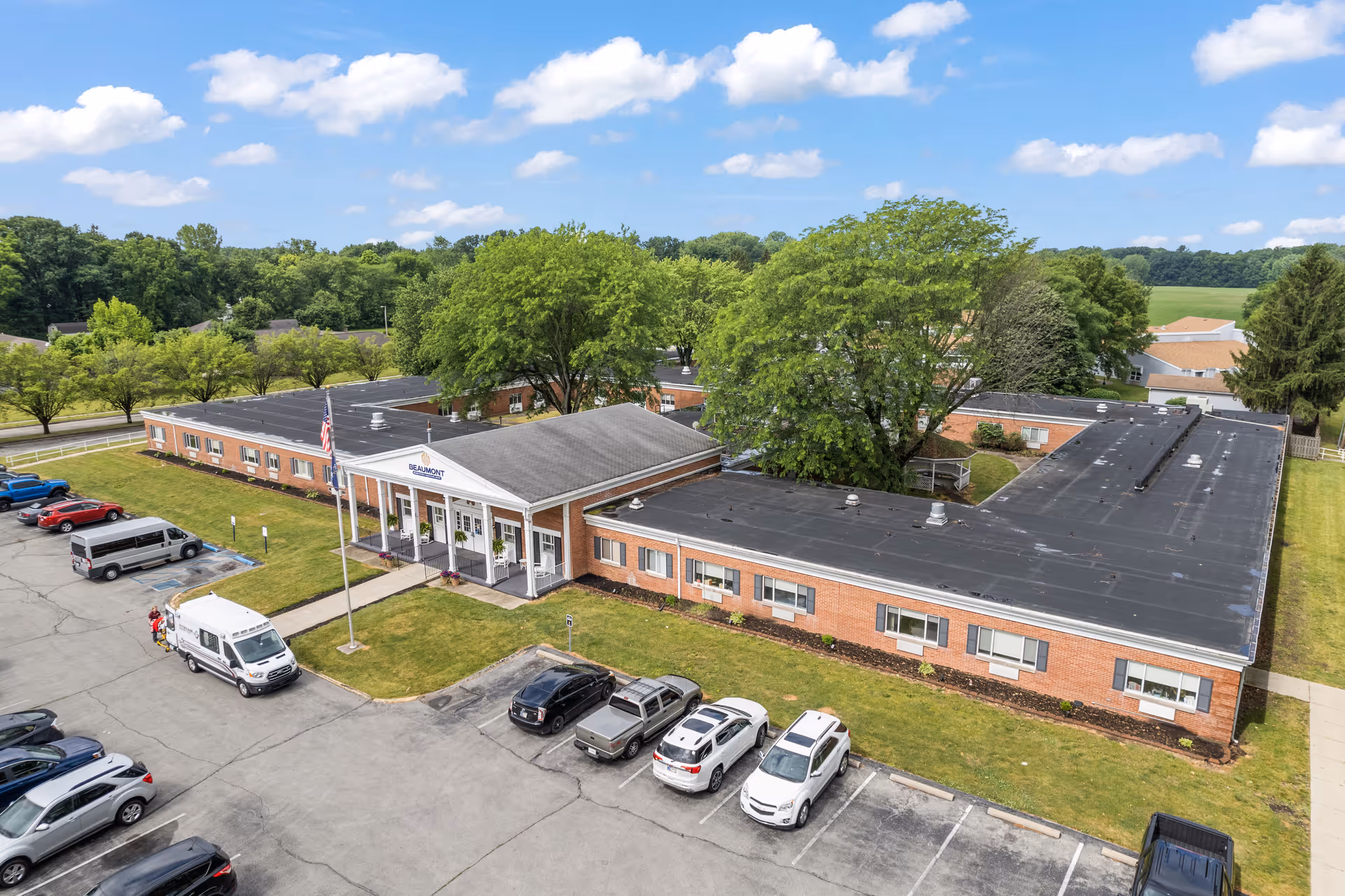 Aerial view of a single-story brick rehabilitation and healthcare center with a parking lot, flagpoles, and surrounding trees.