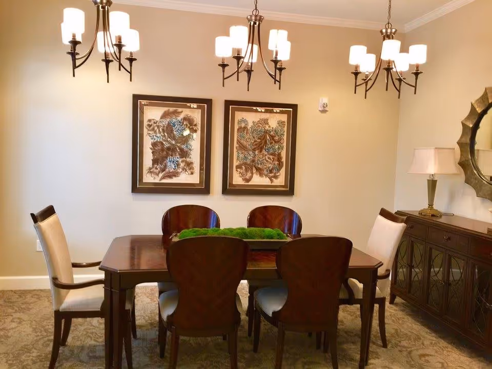 Dining room with a wooden table surrounded by chairs, three chandeliers overhead, framed artwork on the wall, and a sideboard with a lamp.