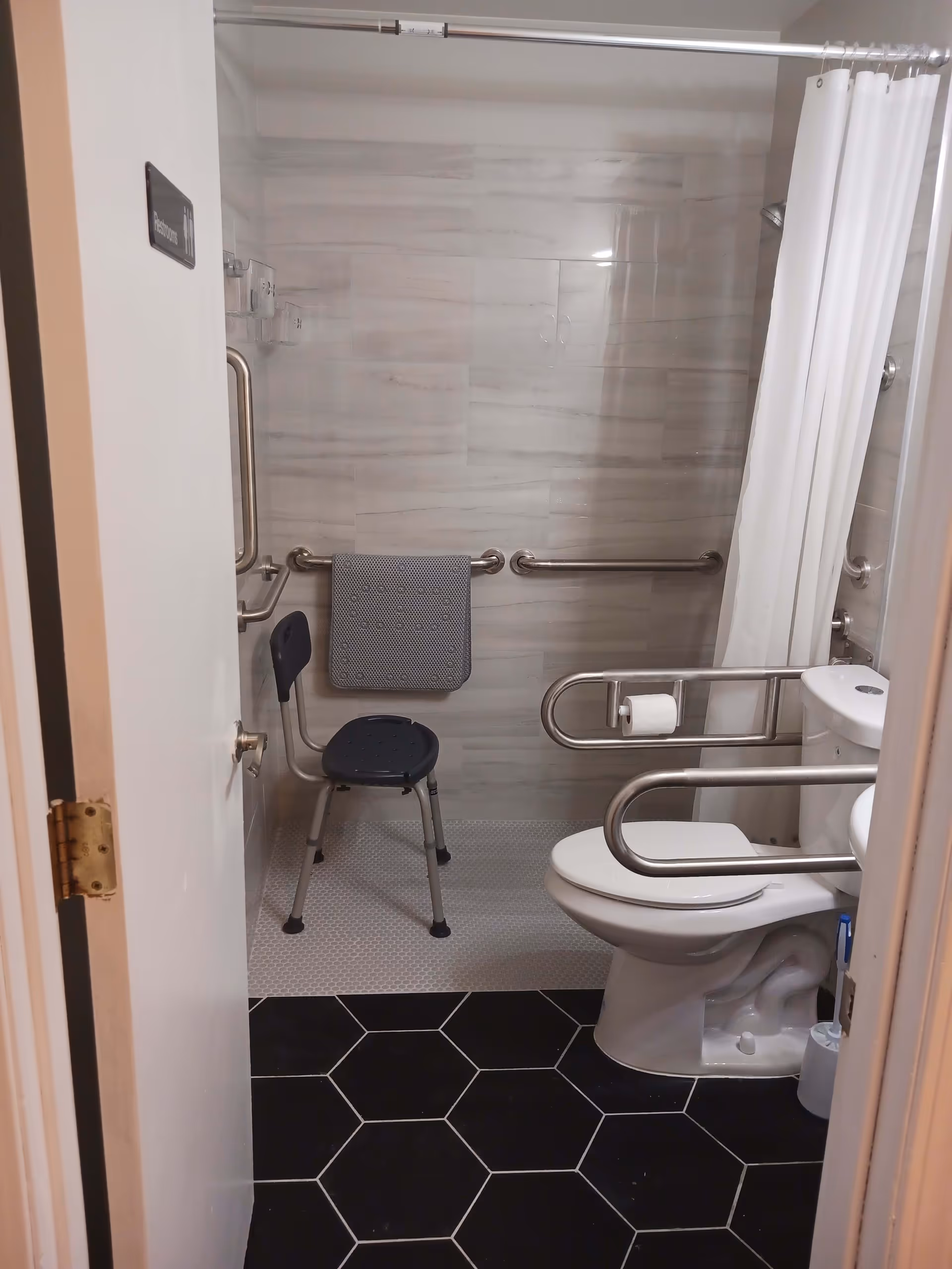 View into an accessible bathroom with a white toilet equipped with safety grab bars, a shower area with a white curtain, a black shower chair, and a towel hanging on a grab bar. The floor has black hexagonal tiles outside the shower and small white tiles inside the shower. The walls are tiled in light gray.
