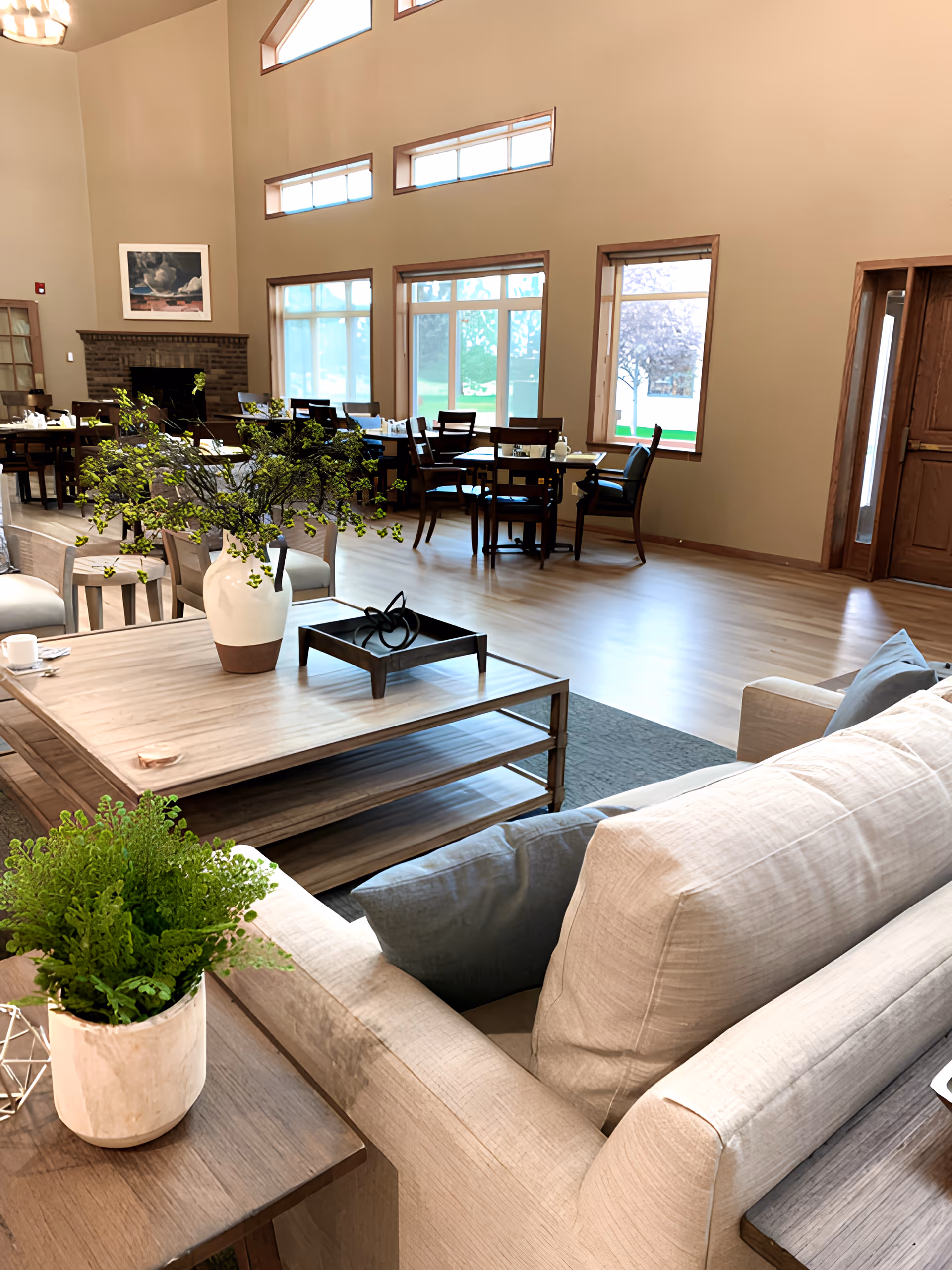 Open, airy senior living common room with sofas and a coffee table in the foreground and dining tables near large windows in the background.