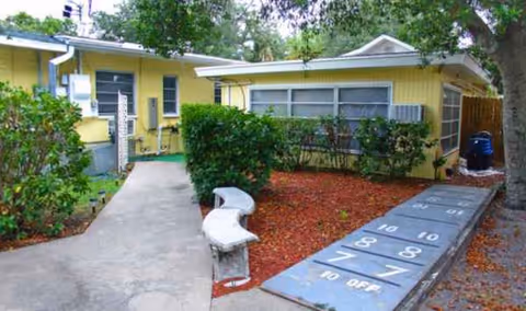Outdoor view of a yellow retirement home with hedges, benches, and a shuffleboard area.