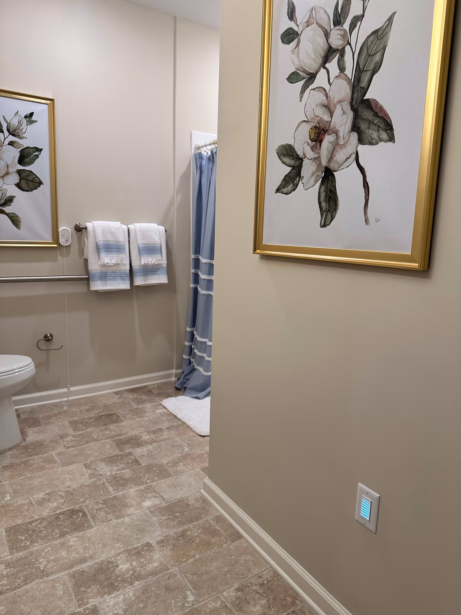 A bathroom with beige walls and tiled floor. There is a toilet on the left side, a towel rack with two white and blue towels, and a blue shower curtain with white trim. Two framed floral paintings with gold frames hang on the walls. A small night light is plugged into a wall outlet near the floor.