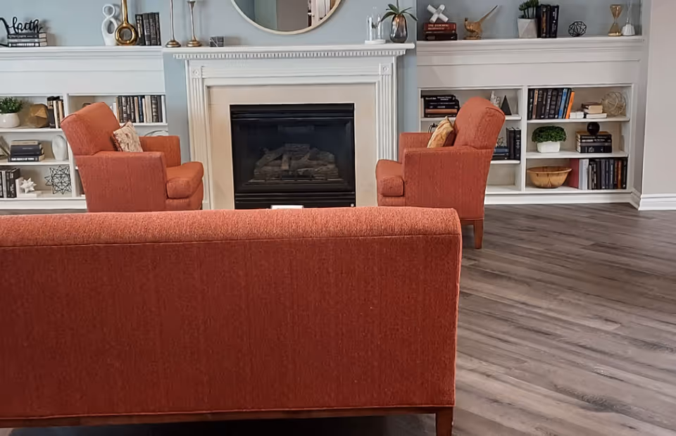 Cozy common living area with a fireplace, built-in bookshelves, and orange sofa and armchairs on wood flooring.
