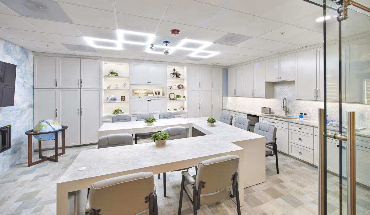 Bright communal activity room with a U-shaped white table surrounded by gray chairs, built-in white cabinets and a kitchenette.