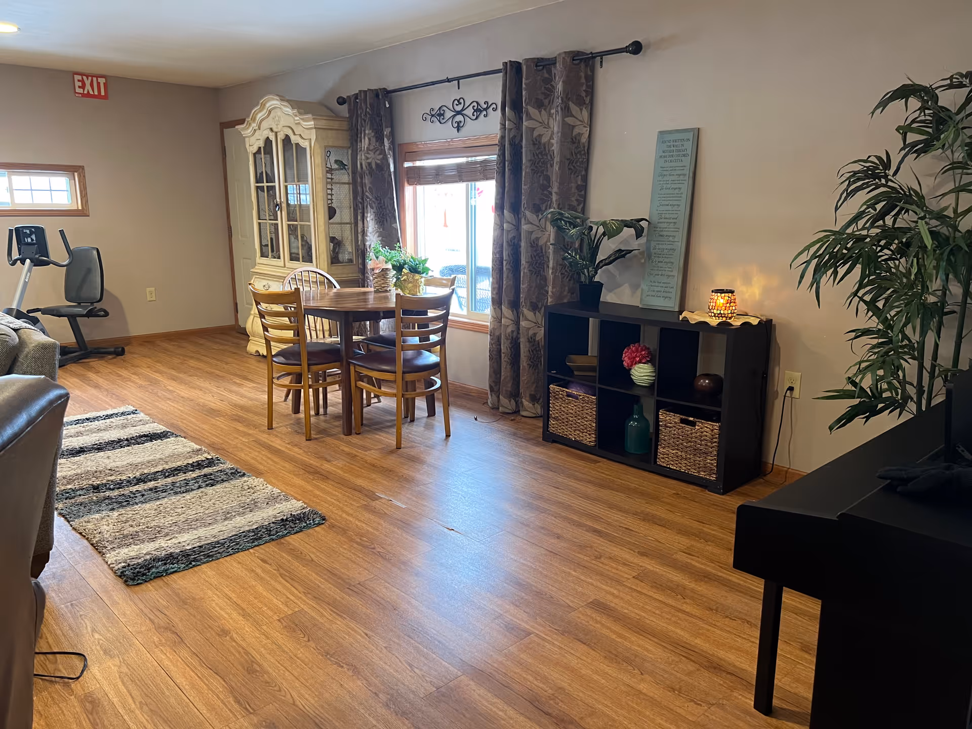 A cozy interior room with wooden flooring featuring a small round dining table with four chairs near a window with floral curtains. There is a decorative cabinet in the corner, a black shelving unit with plants and decorative items, a striped rug, and an exercise bike. The room has neutral-colored walls and a warm, inviting atmosphere.
