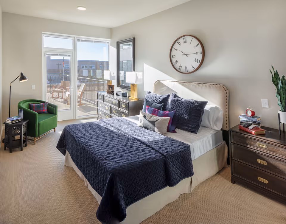 A bright bedroom with a large bed covered in a dark blue quilt and multiple pillows. There is a green armchair with a plaid pillow next to a small side table with books and a lamp. A dresser with two lamps and a mirror is against the wall, and a large clock hangs above the bed. Glass doors lead to an outdoor balcony with two wooden chairs.