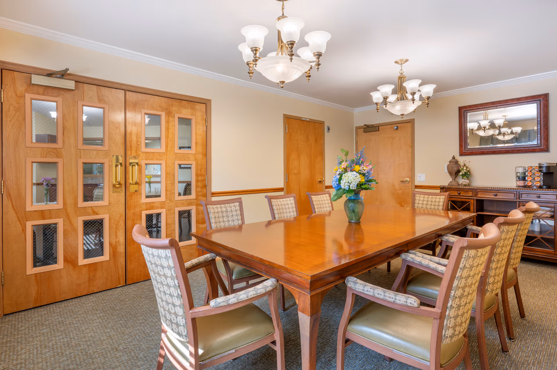 A well-lit dining room with a large wooden table surrounded by eight cushioned chairs. A vase with a colorful flower arrangement is placed in the center of the table. The room features wooden double doors with glass panels, a sideboard with decorative items, a coffee machine, and a large mirror on the wall reflecting the chandelier lighting fixtures above.