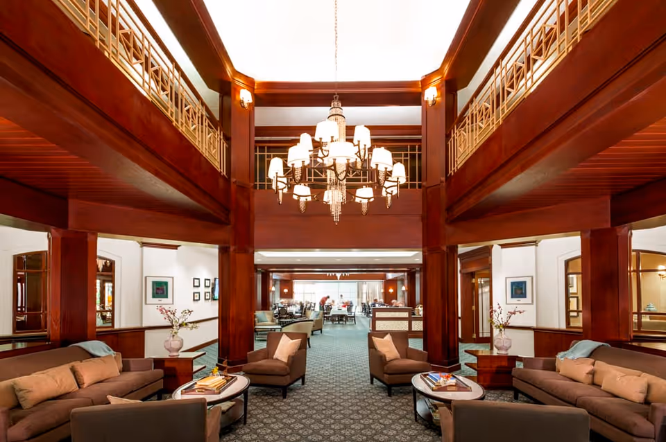 Spacious senior living facility common area with dark wood beams and columns, a large chandelier hanging from the ceiling, multiple seating arrangements including sofas and armchairs with cushions, side tables with decorative vases, and a dining area visible in the background.