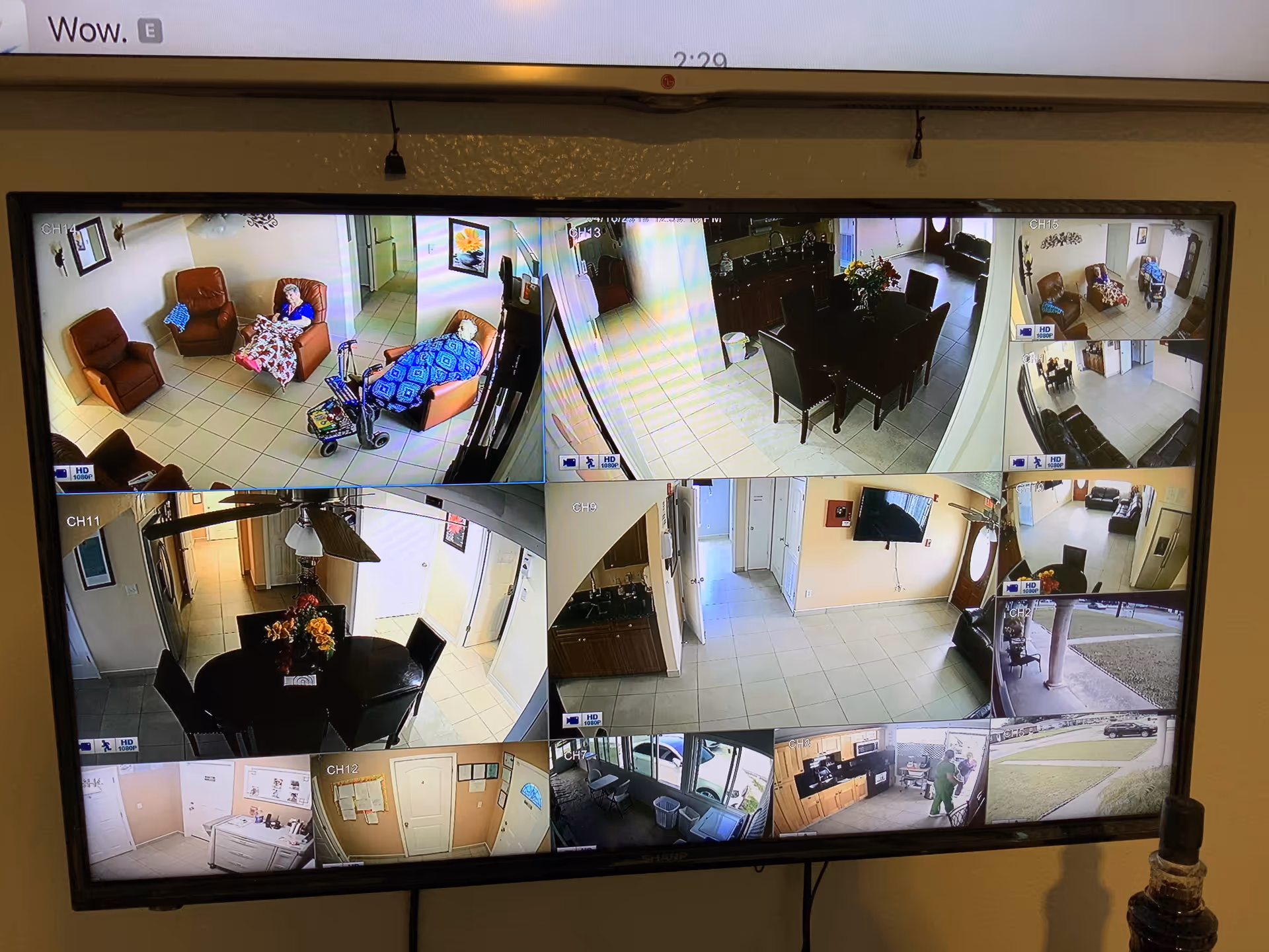 A security monitor screen displaying multiple surveillance camera views of an assisted living facility. Views include common areas with chairs and elderly residents, dining areas with tables and chairs, hallways, an outdoor entrance, and a kitchen area. Some residents are seated and covered with blankets.