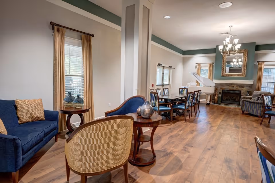A spacious living room area in Town Village Crossing featuring wooden flooring, a blue sofa with patterned cushions, a round wooden side table with decorative vases, several upholstered chairs, a dining table with multiple chairs, a white grand piano near a stone fireplace with a large mirror above it, and windows with beige curtains allowing natural light to enter.
