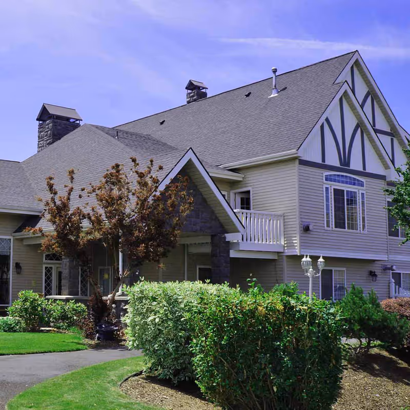 Exterior view of a two-story residential building with beige siding, a gray shingled roof, and decorative timber framing on the upper gable. The building is surrounded by well-maintained landscaping including green bushes, a small tree, and a lawn. There is a paved walkway leading to the entrance and a white balcony on the second floor.