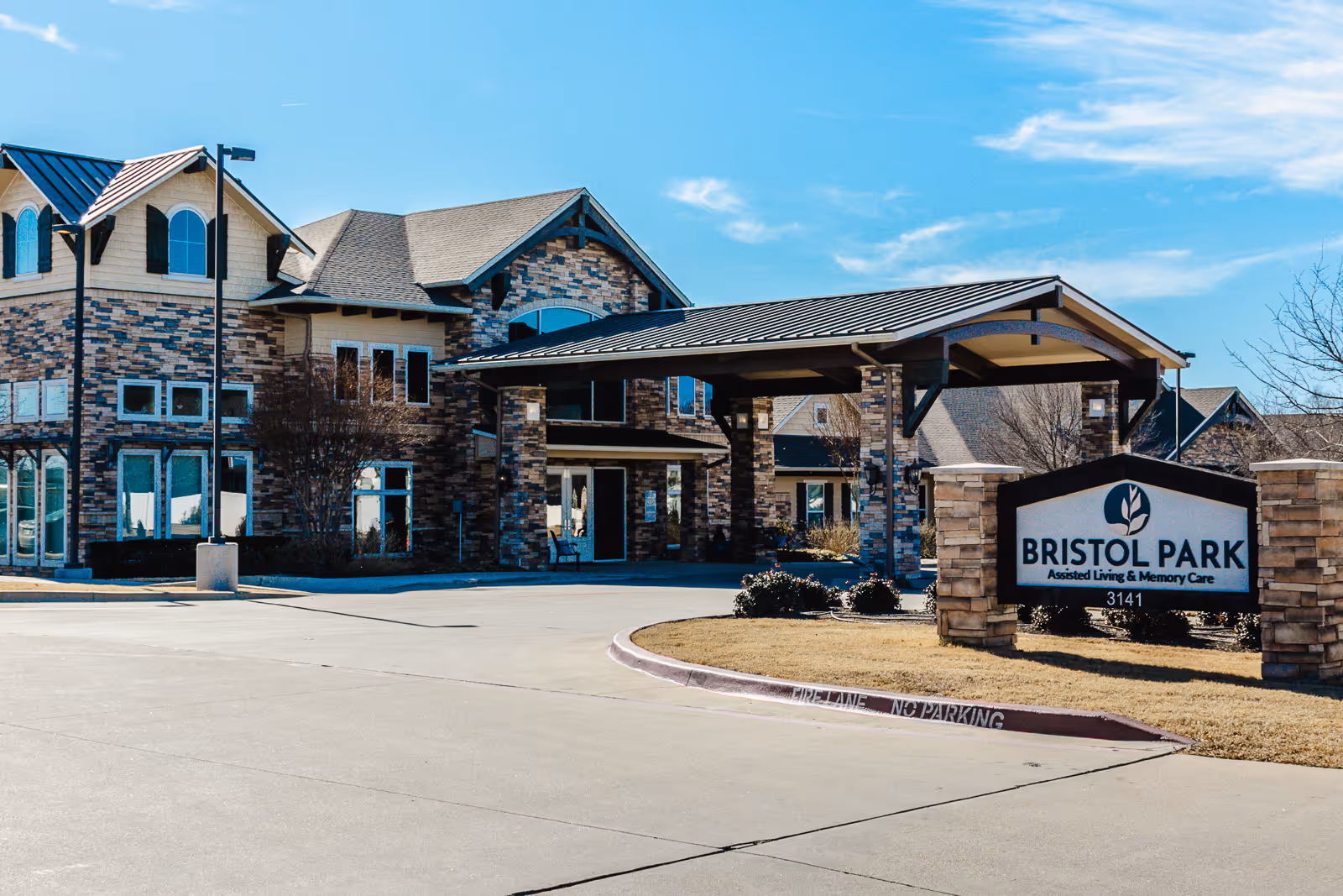 Exterior view of Bristol Park at Eagle Mountain Assisted Living & Memory Care facility showing a large brick building with multiple windows and a covered entrance. A sign in front reads 'Bristol Park Assisted Living & Memory Care 3141'. The sky is clear with a few clouds.
