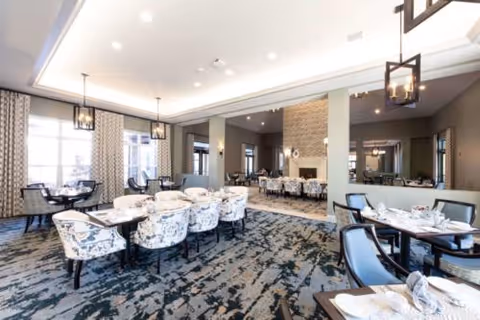 A spacious and elegant dining room with multiple tables set for meals. The room features patterned upholstered chairs, large windows with curtains, modern pendant lighting, and a fireplace in the background. The carpet has a contemporary design in shades of blue and gray.