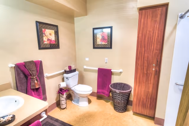 A clean and well-lit bathroom with beige walls and floor. The bathroom features a white toilet with a basket on top, a towel rack with purple and brown towels, a wicker laundry basket, a wooden cabinet, and a sink countertop with a soap dish. Two framed floral pictures hang on the walls above the toilet and towel rack.