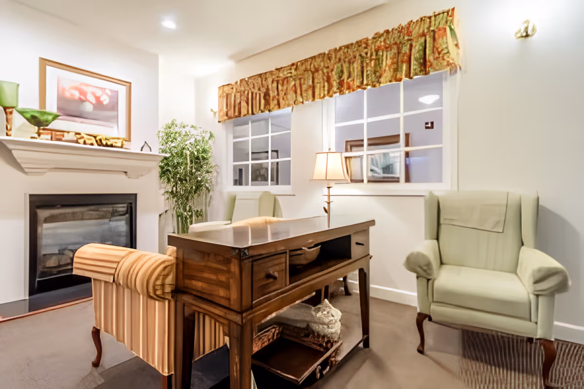 A cozy living room with a fireplace on the left, a wooden table in the center, a striped upholstered armchair behind the table, and a light green armchair to the right. There are two windows with patterned valances, a floor lamp between the windows, and a potted plant near the fireplace.