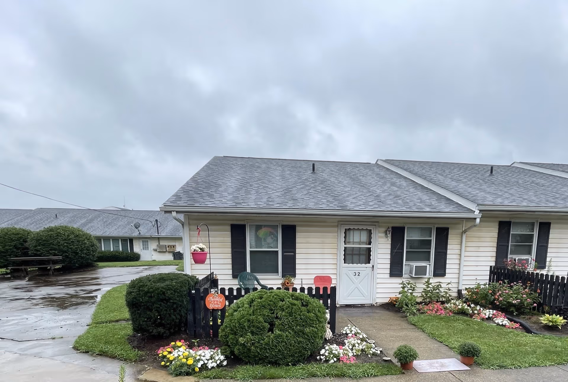 Exterior view of a single-story residential unit with beige siding and a gray shingled roof. The unit has a white door with the number 32 and two windows with black shutters. In front of the unit is a small fenced garden area with green bushes, colorful flowers, and a pink hanging flower pot. The sky is overcast and the ground appears wet from rain.