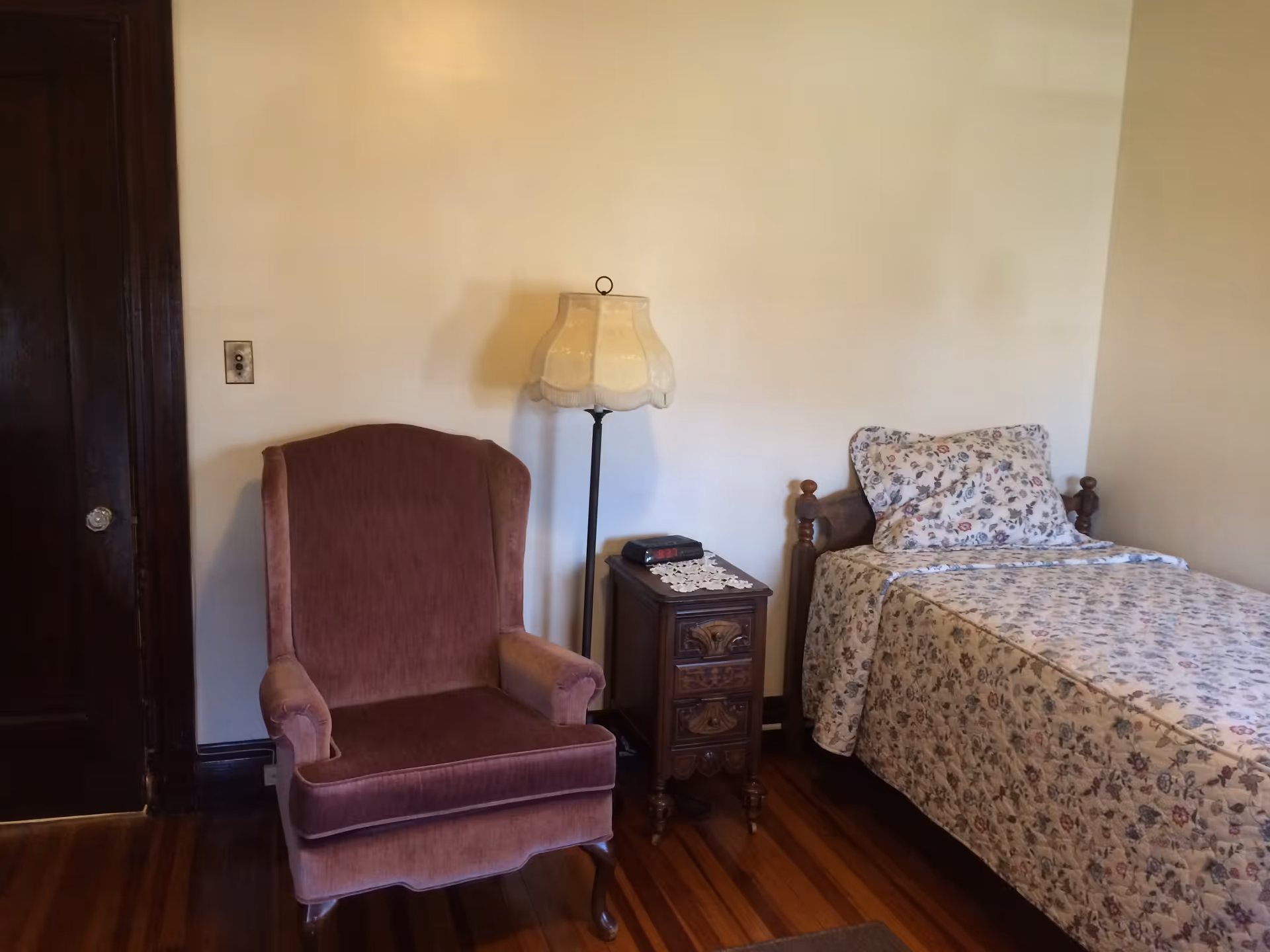 Small bedroom with a floral single bed, wooden nightstand, floor lamp and an upholstered armchair on a hardwood floor.