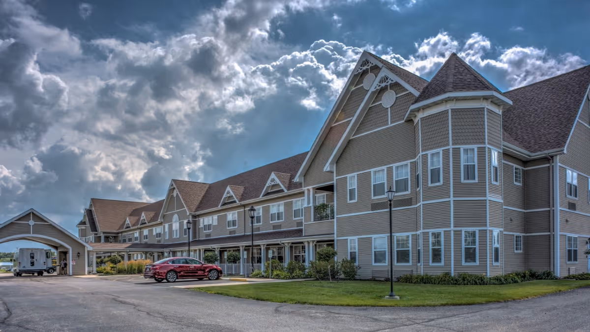 Exterior view of a large, multi-story senior living facility building with beige siding and multiple windows under a partly cloudy sky. There is a driveway with a red car parked and a covered entrance on the left side of the building.