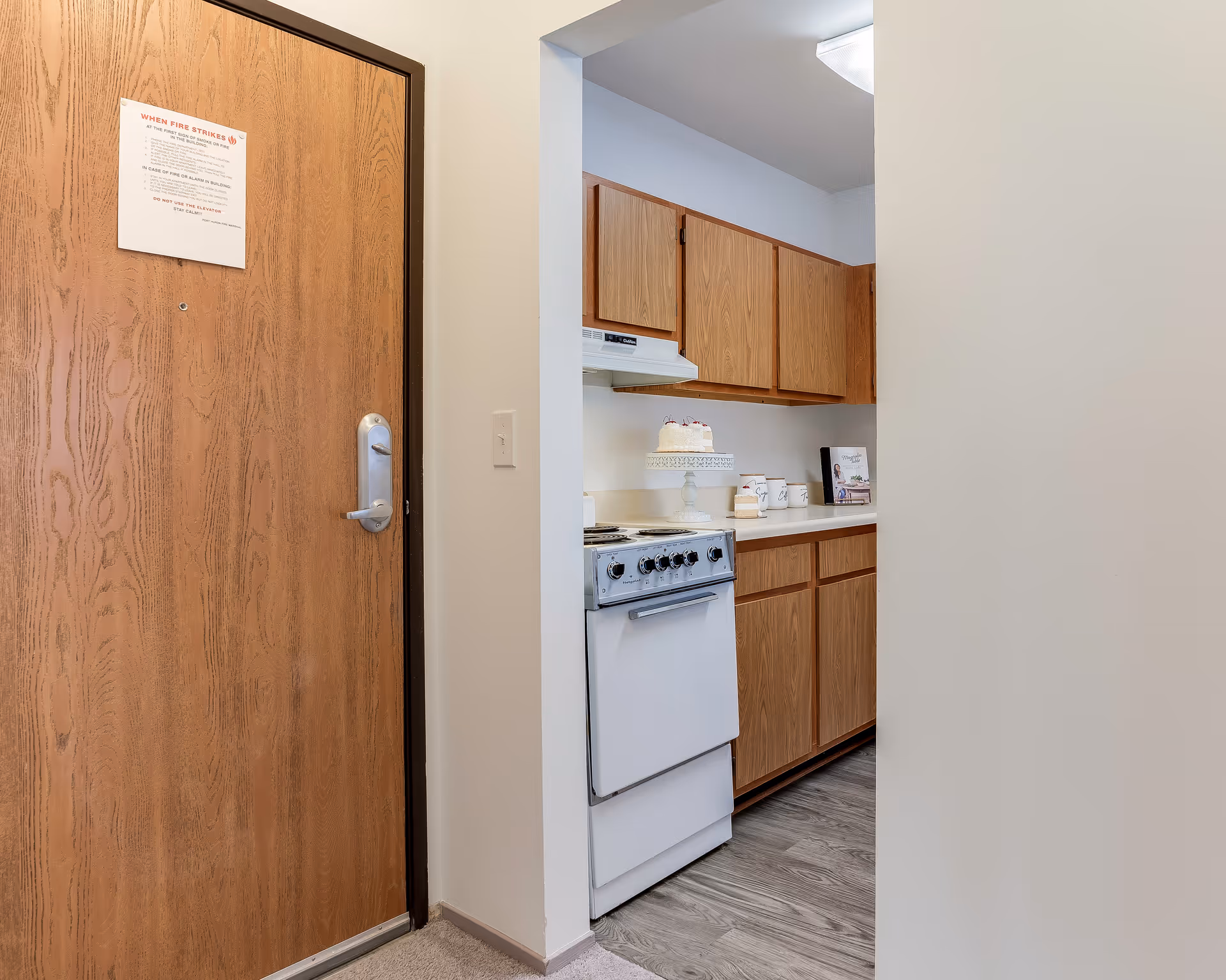 View of a small kitchen area next to a wooden entrance door. The kitchen has wooden cabinets, a white stove with an oven, and a countertop with a cake on a stand and some containers. The floor in the kitchen area is wood-patterned, and the entrance area has carpet.