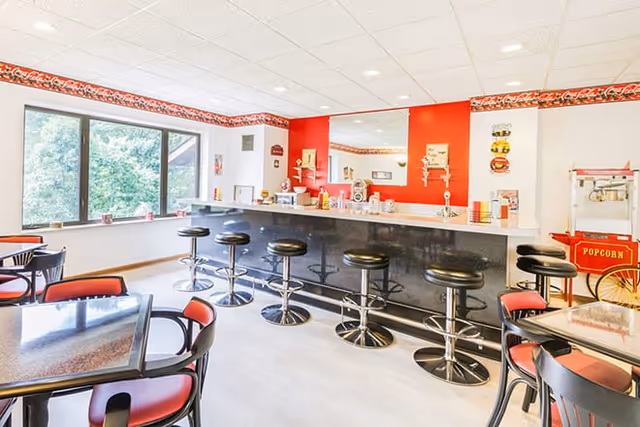 Bright and clean dining area with a long black counter and six black swivel bar stools. Several tables with black chairs featuring red cushions are arranged nearby. A popcorn machine is visible on the right side, and large windows on the left let in natural light with a view of green trees outside. The walls are white with a red accent wall behind the counter and a decorative border near the ceiling.