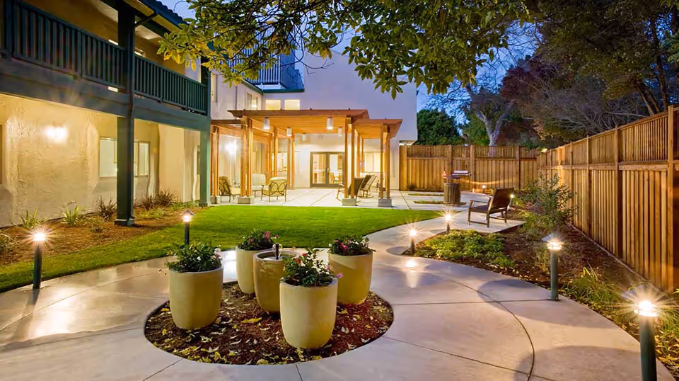 Outdoor courtyard area at dusk with a curved concrete pathway, several large potted plants in the center, small garden lights lining the path, wooden fence on the right, and a covered patio with seating attached to a building.