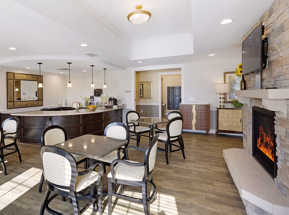 A bright and inviting common area with multiple tables and chairs, a stone fireplace with a TV mounted above it, a countertop with pendant lights hanging overhead, and a decorative mirror on the wall. The room has wooden flooring and modern lighting fixtures.
