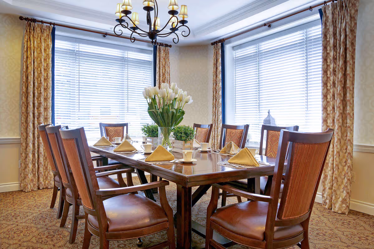 A dining room with a rectangular wooden table set for eight people. The table has yellow folded napkins, white cups and saucers, and a centerpiece with white tulips and green plants. The room has large windows with white blinds and patterned gold curtains, a patterned carpet, and a decorative chandelier hanging above the table.
