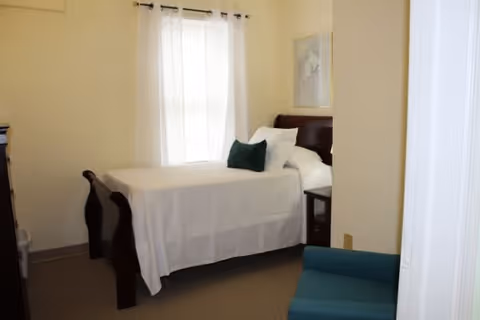 Small furnished bedroom with a single bed dressed in white linens, a green accent pillow, a window with sheer curtains and a teal chair.