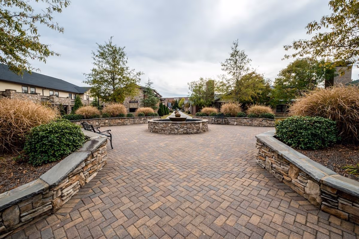 Outdoor courtyard area with a circular stone water fountain in the center, surrounded by stone benches and landscaped bushes and trees. The sky is overcast and buildings with stone facades are visible in the background.