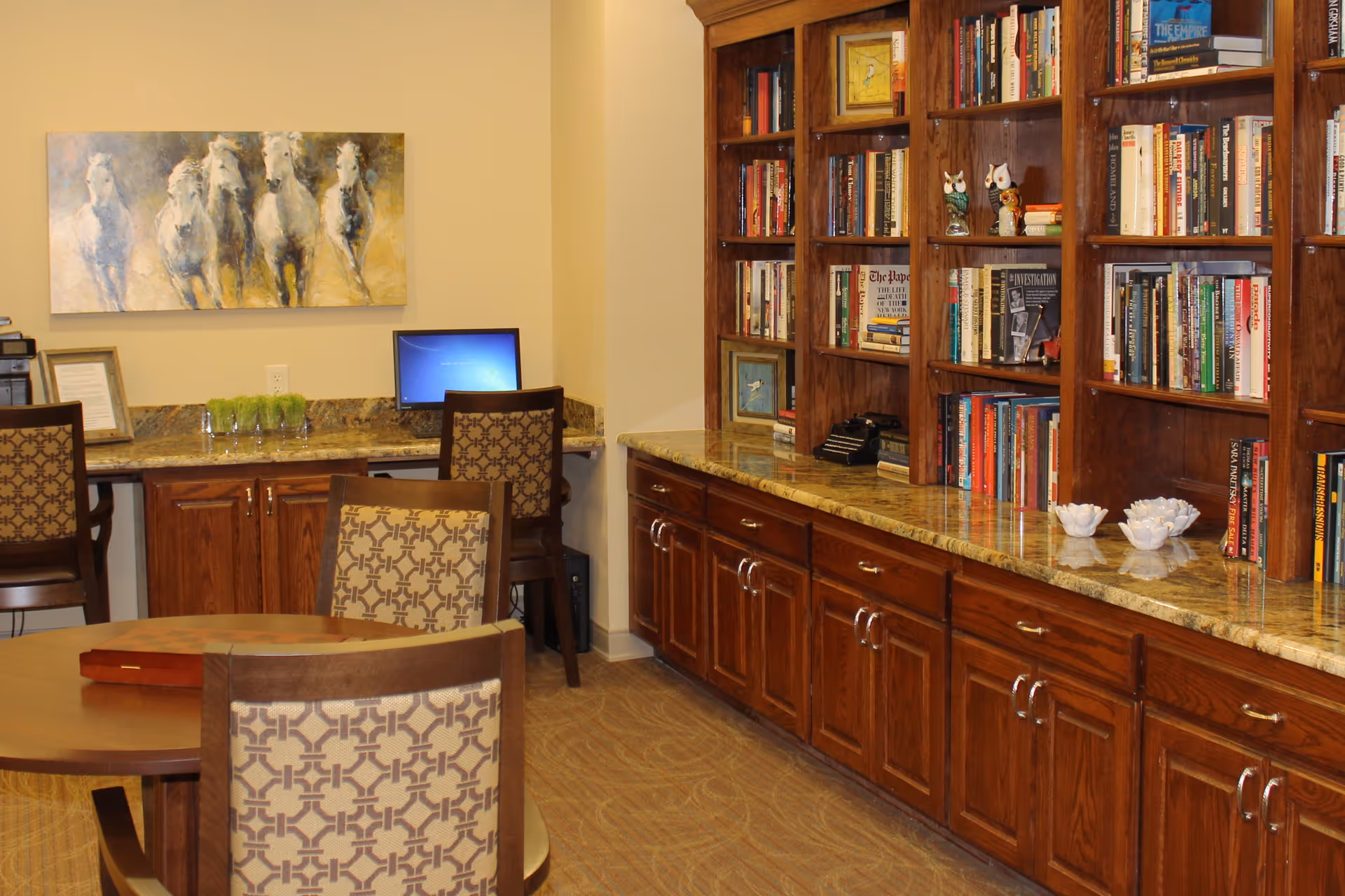 A cozy interior common room featuring built-in wooden bookshelves and cabinets, a long marble countertop, a table with patterned chairs, and a small computer workstation.
