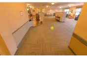 Wide, carpeted common area in a senior living facility with tables, chairs, and seating areas under warm lighting.