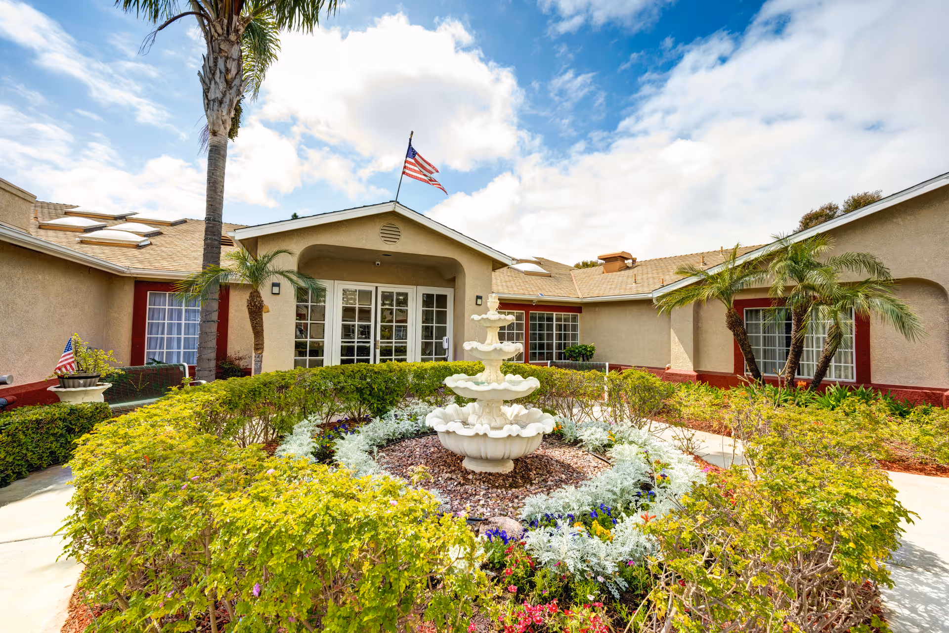 Exterior view of a single-story building with beige walls and red trim, featuring a central entrance with glass double doors. In front of the entrance is a circular garden with a white tiered fountain surrounded by green shrubs and colorful flowers. Palm trees and an American flag on a flagpole are visible under a partly cloudy sky.