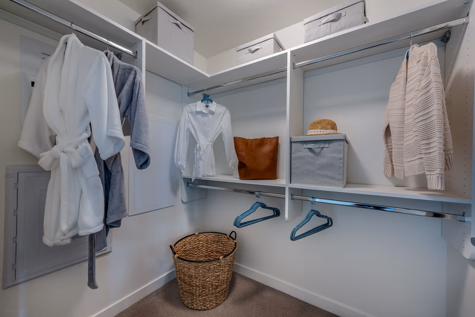 A walk-in closet with white shelving and hanging rods. The closet contains two bathrobes hanging on the left, a white shirt and a brown bag on the middle shelf, a beige sweater hanging on the right, a woven basket on the floor, and two gray storage boxes on the top shelves.