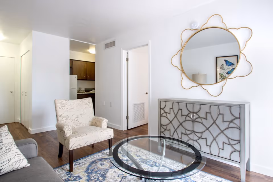 A bright living room with a patterned armchair, a glass coffee table on a blue and white rug, a decorative sideboard with an intricate design, and a round mirror with a gold frame hanging above it. The room has white walls and wooden flooring, with a view into a kitchen area and an open door leading to another room.