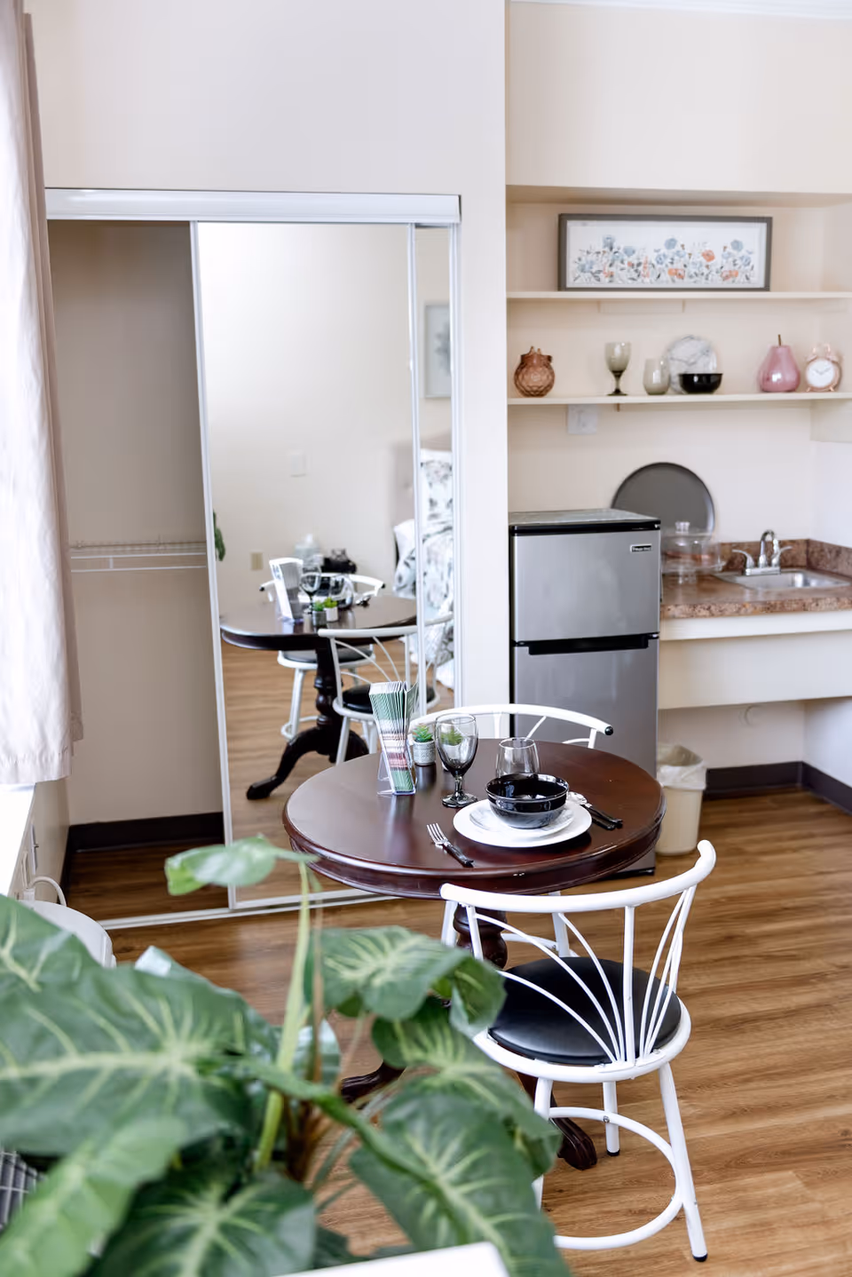 A small dining area with a round wooden table set for two with black bowls, plates, glasses, and cutlery. Two white chairs with black cushions are placed around the table. In the background, there is a small kitchenette with a mini refrigerator, a sink, and shelves holding decorative items. A large mirror with sliding doors reflects part of the room. A green leafy plant is visible in the foreground.