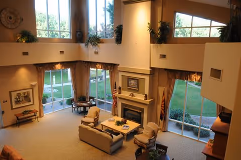 Spacious and well-lit living room area in a senior living community featuring large windows with views of a green outdoor space, comfortable seating including sofas and armchairs arranged around a coffee table, a fireplace with framed artwork above it, and decorative plants placed near the windows and on furniture.